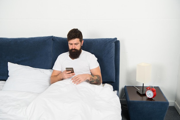 Andare online Uomo barbuto usa lo smartphone a letto Tecnologia per la casa Adozione della tecnologia mobile Tecnologia a banda larga Nuova tecnologia Vita moderna Connessione Internet a casa