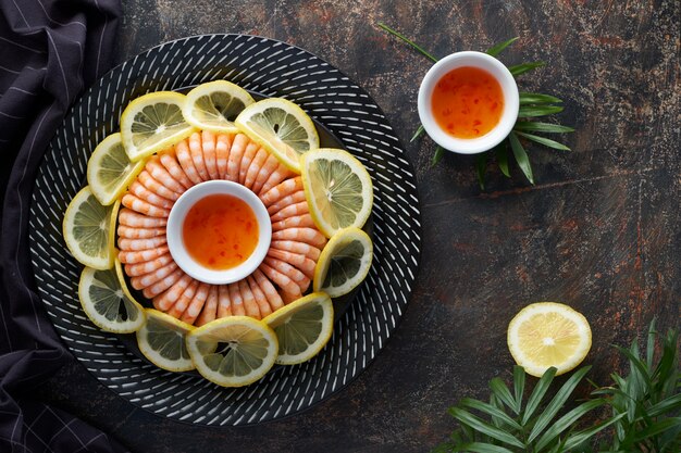 Anello di gustosi gamberi servito su un piatto scuro con limoni e salsa di peperoncino dolce, piatto disteso sul buio