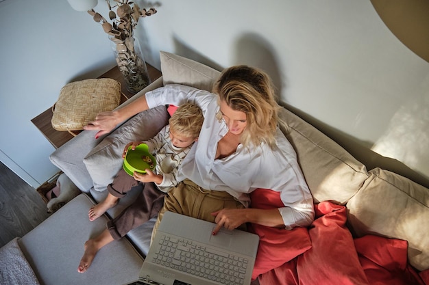 Angolo alto della donna che naviga in netbook e abbraccia il ragazzo che mangia cereali dalla ciotola verde sul divano morbido nel soggiorno di casa