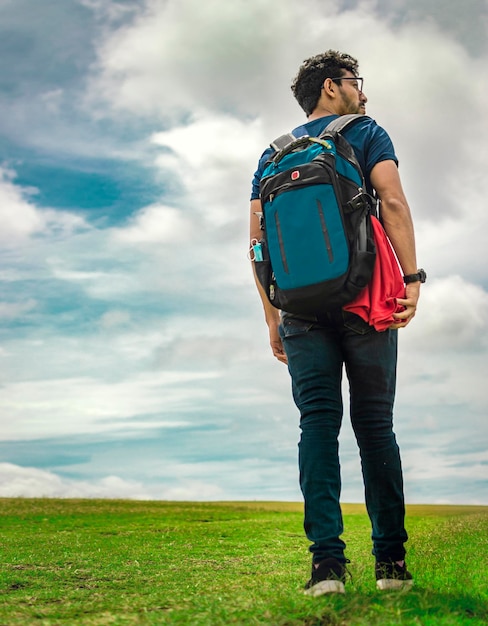 Angolo basso dell'uomo viaggiatore che cammina nel campo Uomo viaggiatore con zaino e sacco a pelo che cammina nella campagna Giovane uomo viaggiatore che cammina nel campo verde Stile di vita dell'uomo avventuroso nel campo