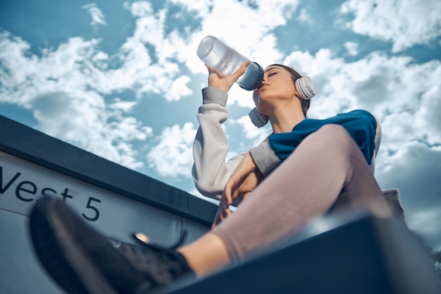 Angolo basso di una giovane donna caucasica moderna e sportiva tranquilla in cuffia che beve acqua all'esterno