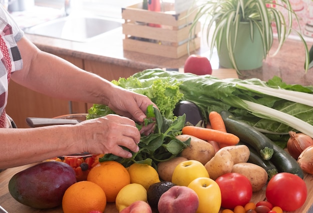 Angolo della cucina e due mani di una donna anziana che lavora per fare una macedonia di frutta fresca. Tavolo in legno con un grande gruppo di frutta e verdura colorata. Mangiare sano