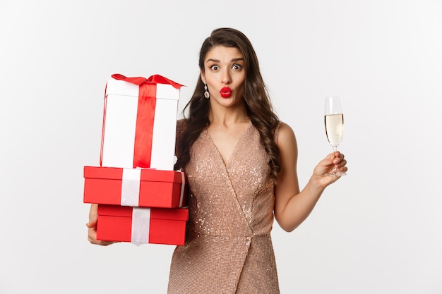 Anno nuovo, Natale e concetto di celebrazione. Donna elegante in abito di lusso e rossetto rosso, con in mano regali e bevendo champagne alla festa, in piedi su sfondo bianco