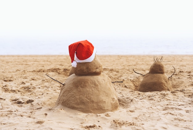 Anno nuovo o buon Natale pupazzo di neve di sabbia con cappello rosso di Babbo Natale sulla spiaggia tropicale dell'oceano