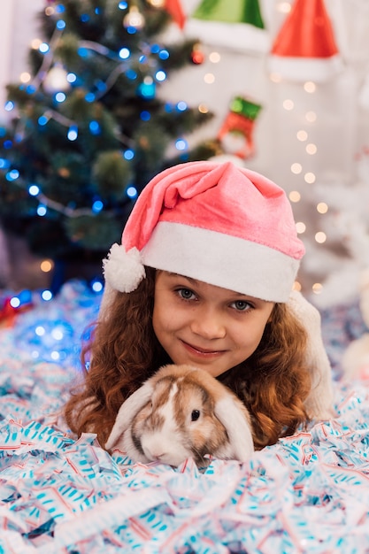 Anno nuovo una ragazza con un cappello rosa da Babbo Natale si trova vicino a un albero di natale e accarezza un coniglio