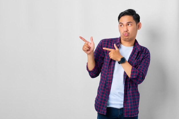 Annoiato giovane asiatico in camicia a quadri che punta il dito contro il banner pubblicitario dello spazio di copia isolato su sfondo bianco