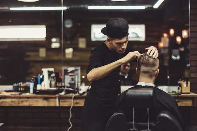 Anonimo barbiere elegante con tatuaggi che tagliano i capelli del cliente maschio sulla sedia