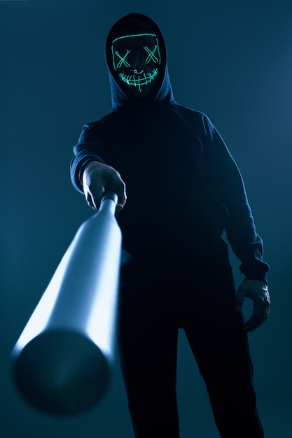 Anonimo uomo criminale con mazza da baseball in una felpa con cappuccio nera e maschera al neon