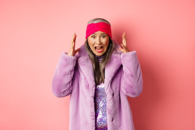 Ansiosa donna anziana asiatica in elegante cappotto di pelliccia sintetica che urla, stringe la mano e grida allarmata e preoccupata alla telecamera, in piedi su sfondo rosa