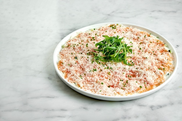 Antipasto italiano classico - carpaccio di salmone con rucola e parmigiano, servito su un piatto bianco su un tavolo di marmo. Salmone a fette sottili. Ristorante di pesce.