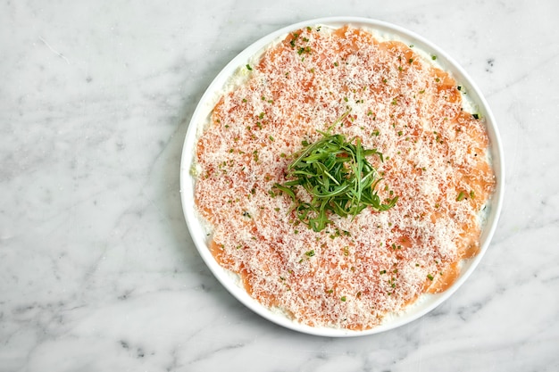 Antipasto italiano classico - carpaccio di salmone con rucola e parmigiano, servito su un piatto bianco su un tavolo di marmo. Salmone a fette sottili. Ristorante di pesce.