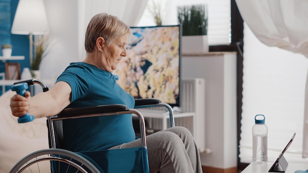 Anziana con disabilità che utilizza manubri per esercitarsi e guardare video di allenamento su tablet digitale. Persona in pensione seduta su una sedia a rotelle, che si allena con i pesi e guarda la lezione online