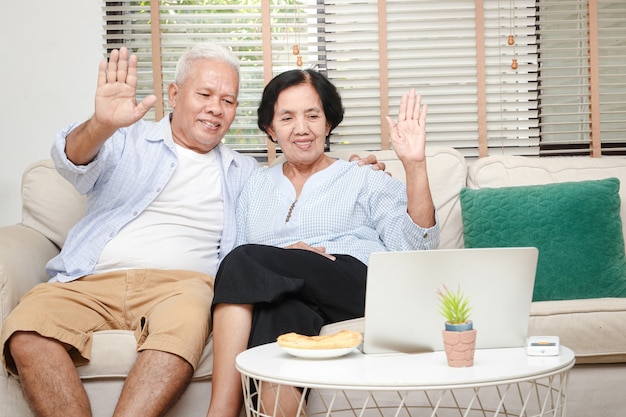 anziana coppia asiatica seduta in soggiorno Alza la mano per salutare figli e nipoti Via video online sul laptop