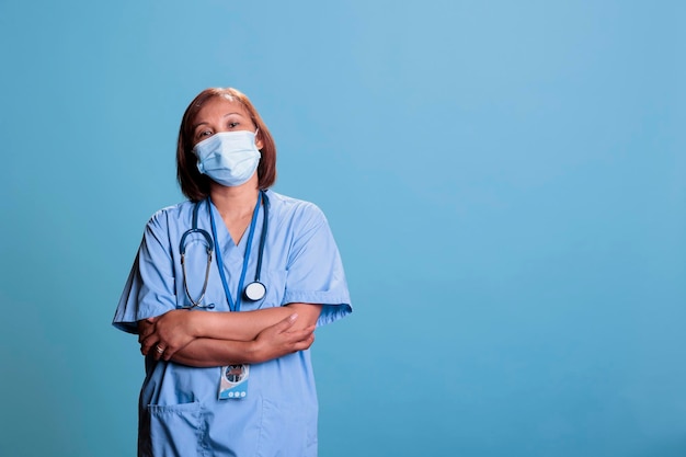 Anziana infermiera fiduciosa in piedi in studio con sfondo blu con il braccio incrociato durante l'appuntamento per la visita di controllo medico. Assistente che indossa una maschera facciale contro il coronavirus, sistema sanitario