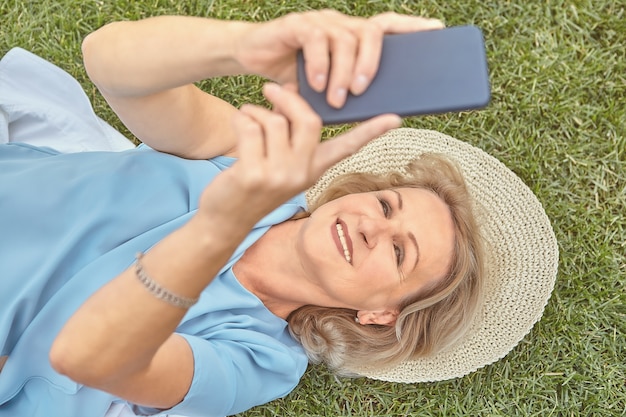 Anziana signora piuttosto caucasica di circa 62 anni è sdraiata sull'erba all'aperto con cappello e smartphone in mano e sorridente.