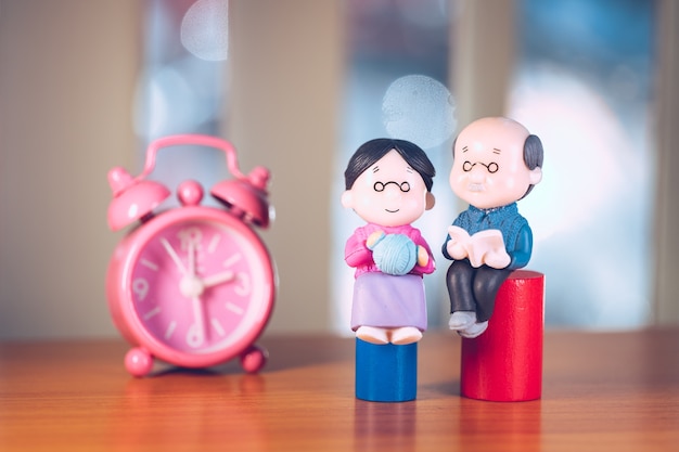 Anziani in miniatura, nonno e nonna seduti sullo sfondo dell'orologio rosa usando come concetto di famiglia e assicurazione