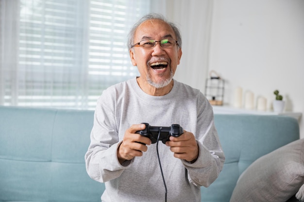 Anziano anziano asiatico che si diverte a tenere il joystick giocando a un videogioco a casa