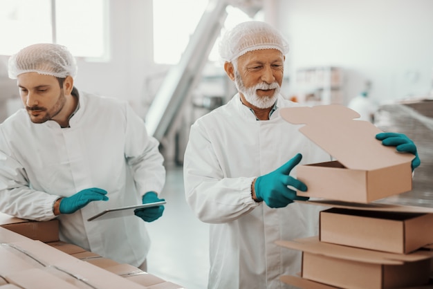 Anziano di lavoro caucasico in uniforme bianca che mette e prepara le scatole mentre sorridente supervisore che tiene compressa e caselle di controllo. Interno della pianta alimentare.