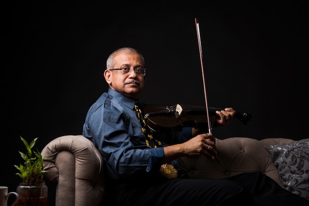 Anziano indiano asiatico bell'uomo che suona o posa con uno strumento musicale a corda come chitarra o violino, in piedi su sfondo nero o seduto sul divano o sul divano