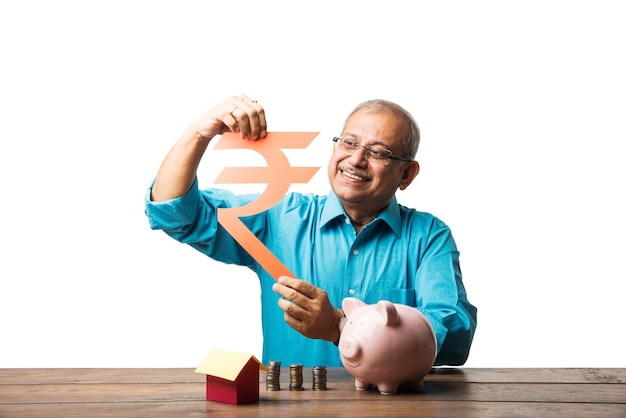 Anziano uomo asiatico indiano e concetto di risparmio - con salvadanaio, banconote, modello di casa 3d, ventaglio di soldi, mucchio di monete. seduto a tavola