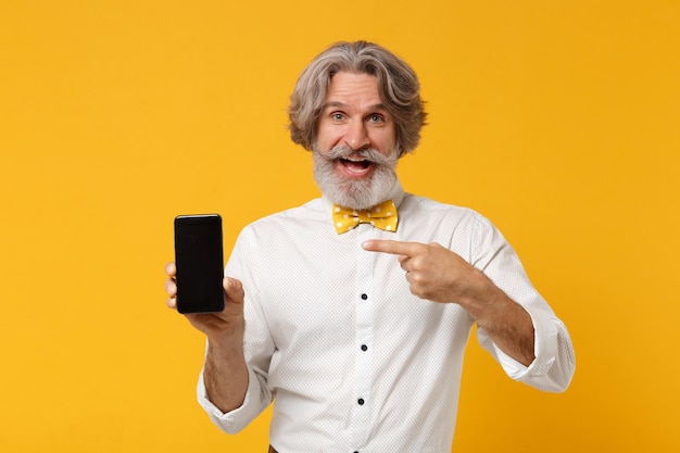 Anziano uomo barbuto con baffi dai capelli grigi in cravatta a farfalla isolata su sfondo giallo. Concetto di stile di vita delle persone. Mock up spazio di copia. Puntare il dito indice sul telefono cellulare con schermo vuoto vuoto.