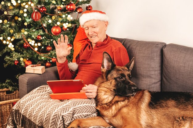 Anziano uomo caucasico in cappello della Santa con tablet seduto su un divano vicino a un albero di Natale con confezione regalo e cane pastore tedesco in chat con i parenti online. Autoisolamento, atmosfera vacanziera.