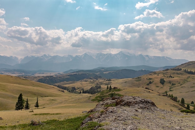 Aperta campagna con un paesaggio vasto e spazioso con un prato verde, molti alberi e un grande acciotolato davanti. Sfondo di alte montagne rocciose sotto un cielo blu con poche nuvole.