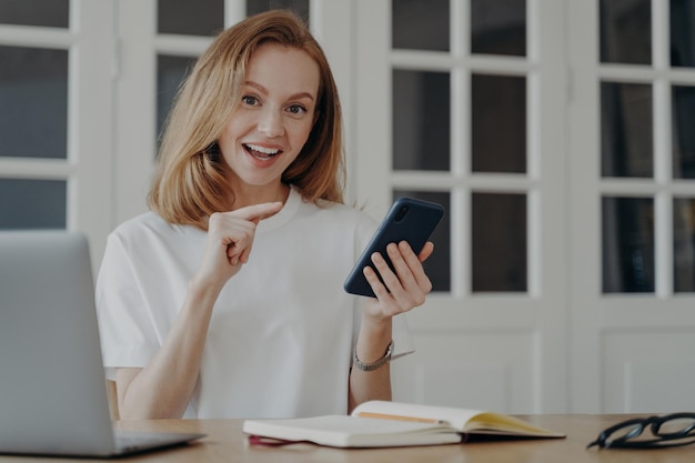 App mobili pubblicitarie femminili eccitate che puntano il dito sullo smartphone seduto alla scrivania con il laptop