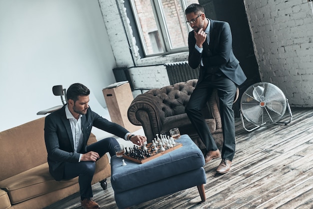 Appassionato di scacchi. Giovani uomini premurosi in abiti completi che giocano a scacchi seduti in casa