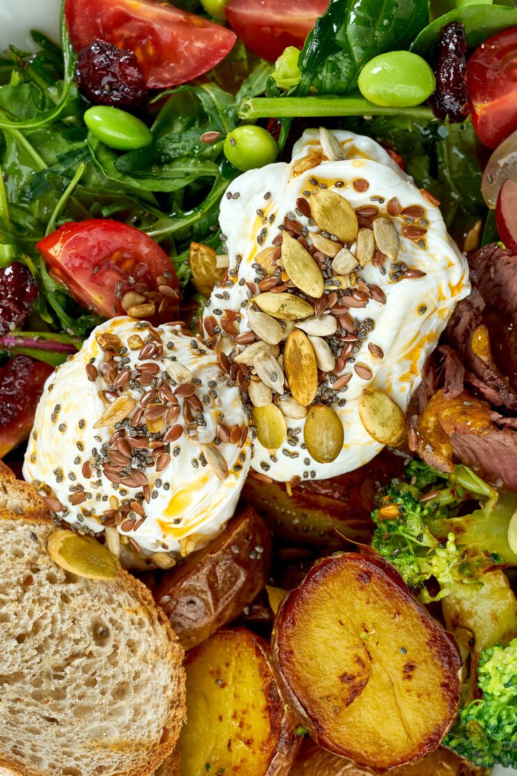 Foto appetitosa e ricca colazione con uovo in camicia, patate, pancetta, uva, insalata mista e pane di segale in una ciotola bianca sul tavolo di legno
