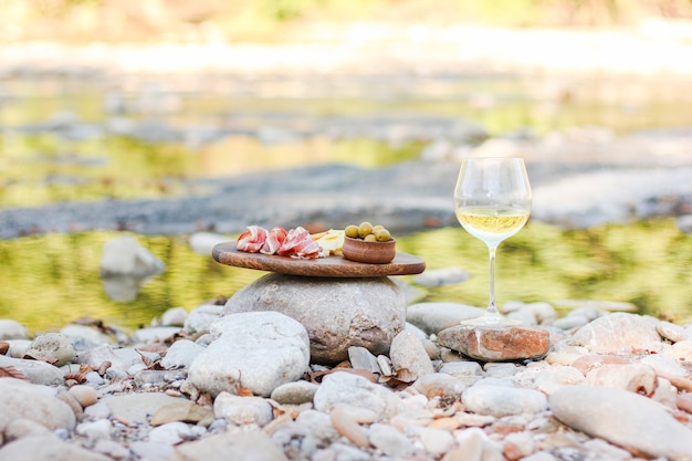 Appetitosi spuntini deliziosi di carne e formaggio su una tavola di legno e un bicchiere di vino stanno sulla riva del fiume durante un picnic