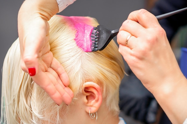 Applicare la tintura rosa con il pennello sui capelli bianchi di una giovane donna bionda in un parrucchiere
