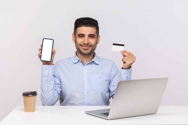 Applicazione bancaria online Uomo d'affari felice seduto sul posto di lavoro dell'ufficio con carta di credito e telefono cellulare con display mock up vuoto spazio libero per la pubblicità girato in studio al coperto isolato