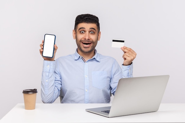 Applicazione bancaria online. Uomo d'affari scioccato sorpreso seduto sul posto di lavoro dell'ufficio, in possesso di carta di credito e telefono cellulare con display mock up vuoto, spazio libero per la pubblicità. girato in studio isolato