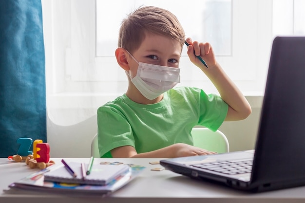 Apprendimento a distanza, istruzione online. Distanza sociale, autoisolamento durante la quarantena. Bambino in età prescolare o scolaro nella mascherina medica che studia a casa con il computer portatile, facendo i compiti per la scuola di sviluppo