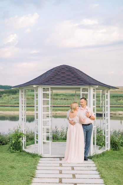 Appuntamento romantico sulla riva dello stagno. Felice coppia matura, uomo e donna in abiti eleganti, in piedi vicino a gazebo in legno sulla riva del bellissimo lago