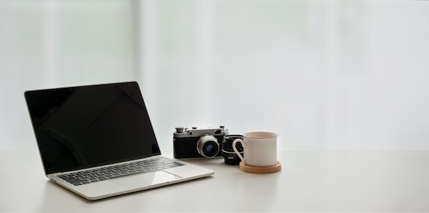 Apra il computer portatile con la tazza di caffè e la macchina fotografica d'annata sulla tavola bianca