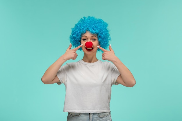 April Fools Day ragazza carina dita puntate sul viso con naso rosso in un costume da clown capelli blu