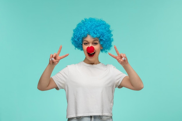 April Fools Day ragazza felice che mostra il segno di pace a bocca aperta con il naso rosso in costume da clown capelli blu