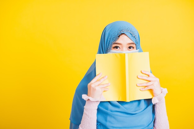 Arabo musulmano asiatico, Felice bella giovane donna religiosa indossa velo hijab e maschera per il viso per prevenire il coronavirus lei studente tiene i libri vicino al viso, isolato su sfondo giallo, Ritorno al college