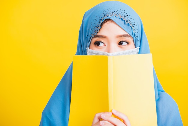 Arabo musulmano asiatico, Felice bella giovane donna religiosa indossa velo hijab e maschera per il viso per prevenire il coronavirus lei studente tiene i libri vicino al viso, isolato su sfondo giallo, Ritorno al college