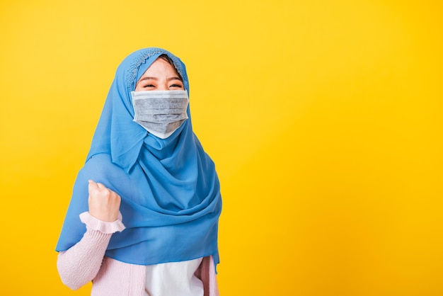 Arabo musulmano asiatico, Ritratto di felice bella giovane donna Islam indossare velo hijab e maschera facciale proteggere alzare le mani felice eccitato allegro dopo essersi ripreso dalla malattia coronavirus isolato giallo