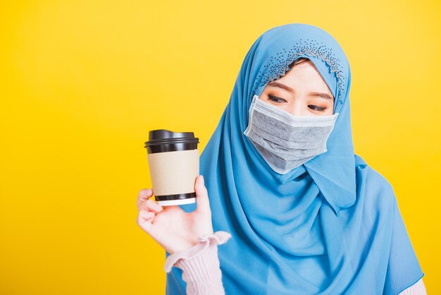 Arabo musulmano asiatico, Ritratto di felice bella giovane donna Islam religioso indossa velo hijab e maschera facciale protegge lei mette in quarantena malattia coronavirus tenere caffè tazza di carta isolato sfondo giallo