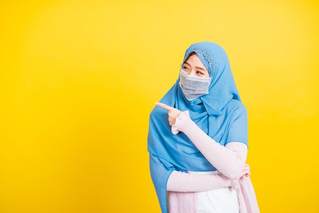 Arabo musulmano asiatico, Ritratto di felice bella giovane donna religiosa indossa velo hijab e maschera facciale protettiva per prevenire il coronavirus lei punta il dito lateralmente nello spazio, isolato su sfondo giallo