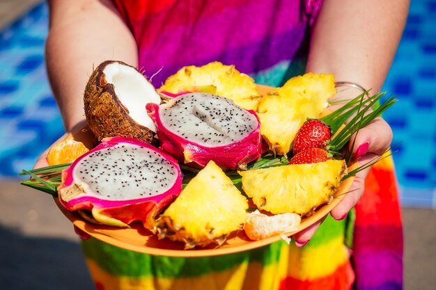 Arancia, cocco, melograno, dragonfruit, ananas su un piatto nelle mani di una donna in costume da bagno sullo sfondo della piscina. disintossicazione