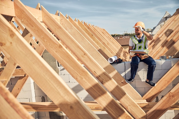 Architetto ambizioso che tocca il suo casco protettivo mentre tiene in mano un blocco per appunti e si siede in un cantiere edile