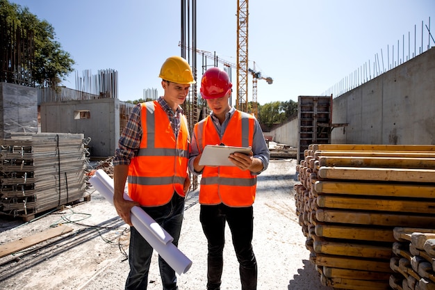 Architetto civile e direttore dei lavori vestiti con giubbotti e caschi da lavoro arancioni discutono di un progetto di costruzione sul tablet mobile nel cantiere aperto accanto alla gru. .