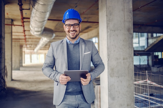 Architetto con la barba lunga caucasico allegro bello in vestito, con il casco sulla testa e tablet nelle mani in piedi in edificio nel processo di costruzione