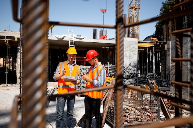 Architetto e ingegnere strutturale in giubbotti e caschi da lavoro arancioni discutono di un progetto di costruzione in un cantiere all'aperto con molti telai in acciaio. .