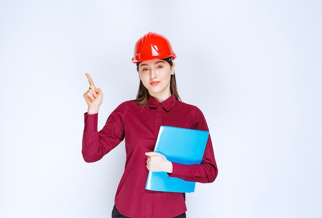 Architetto femminile in casco rosso che tiene cartella blu con documenti e puntamento.